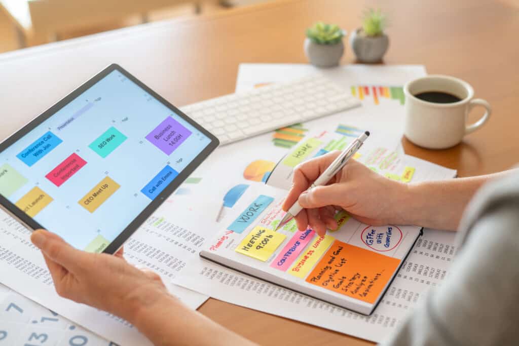 Business professional using a digital calendar on a tablet while writing in a planner, organizing work tasks and scheduling professional activities.